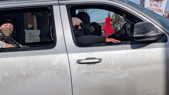 Masked ICE agents, one showing a gun, ride through a protest in a large gray vehicle.